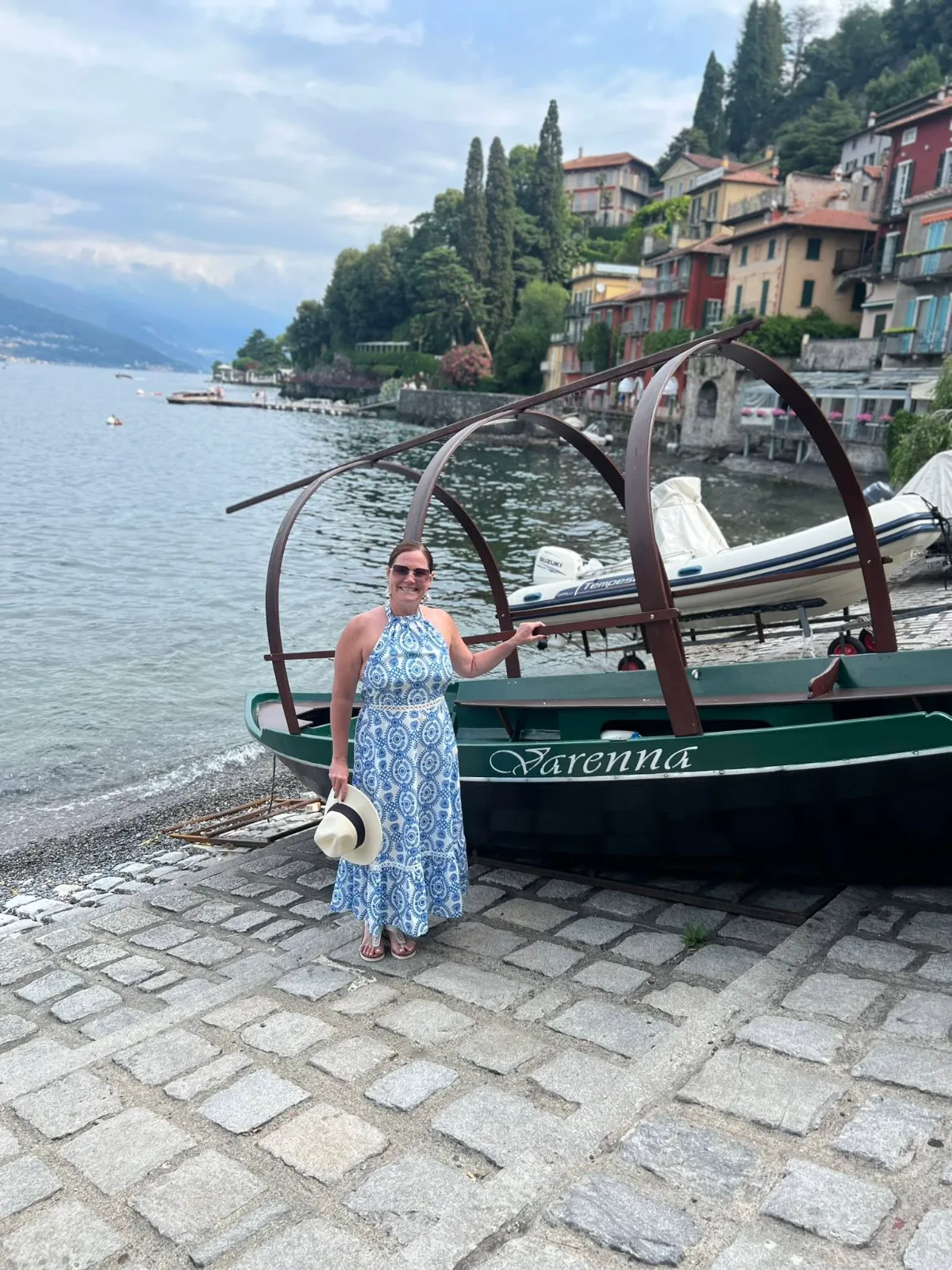 A smiling woman in a patterned sundress stands beside a traditional boat on the waterfront in Varenna, Lake Como.