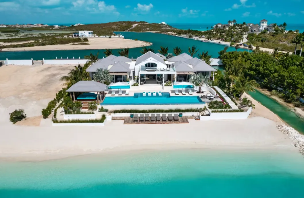 A modern white villa with an infinity pool overlooks a white sand beach and turquoise coastal water.