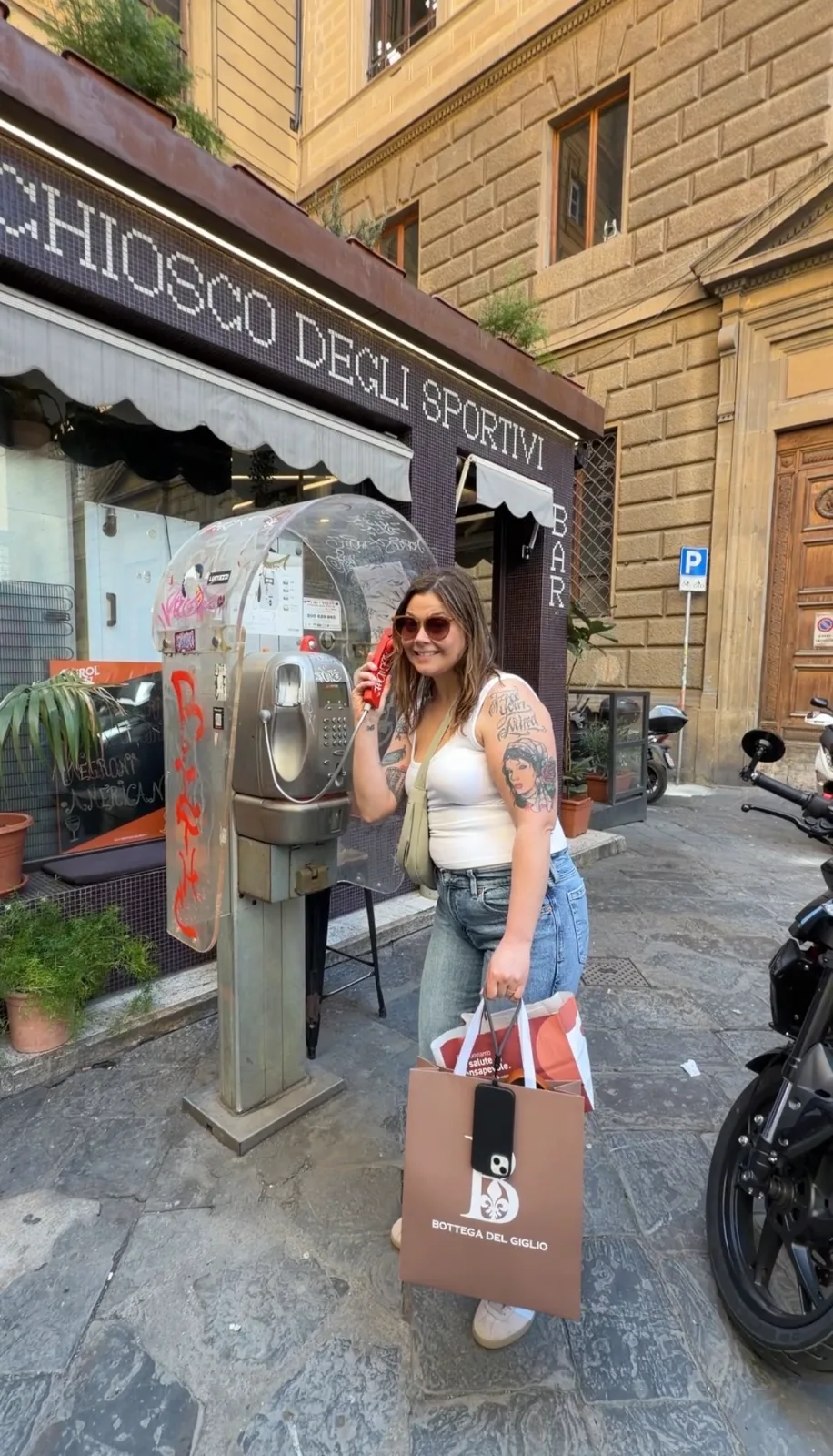 A smiling woman holding shopping bags poses with a vintage payphone outside a traditional café along an Italian street.