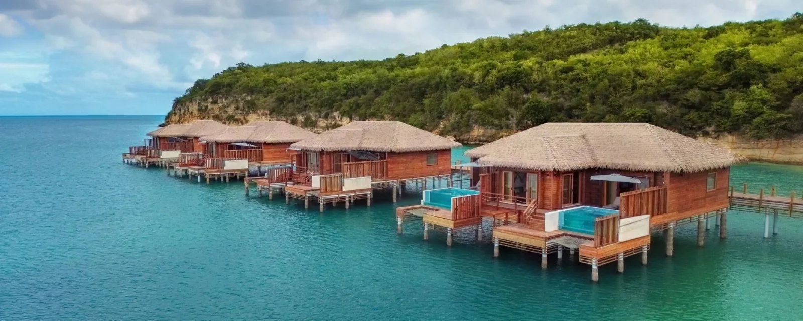 Overwater wooden bungalows with thatched roofs and private plunge pools sit above turquoise waters along a lush coastline in Antigua.