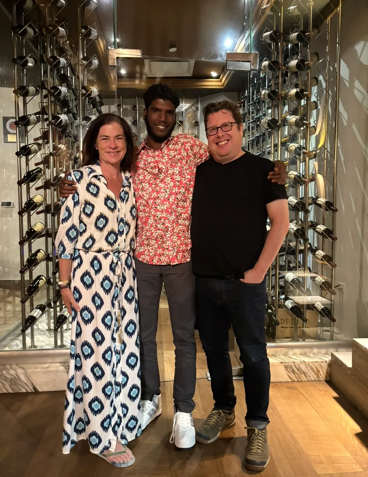 Three smiling adults pose together in front of a modern glass-enclosed wine cellar filled with bottles.