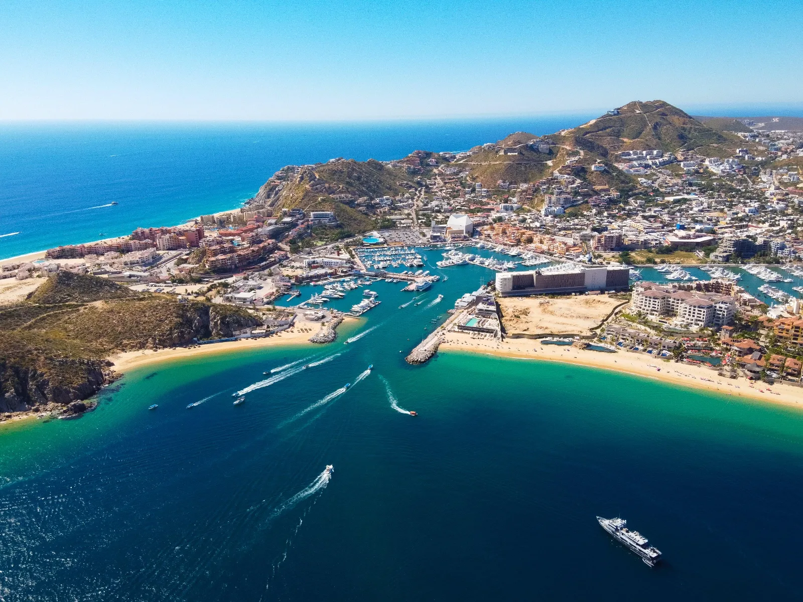 Aerial view of boats navigating turquoise waters into the marina at Cabo San Lucas, Mexico, bordered by sandy beaches and rugged hills.