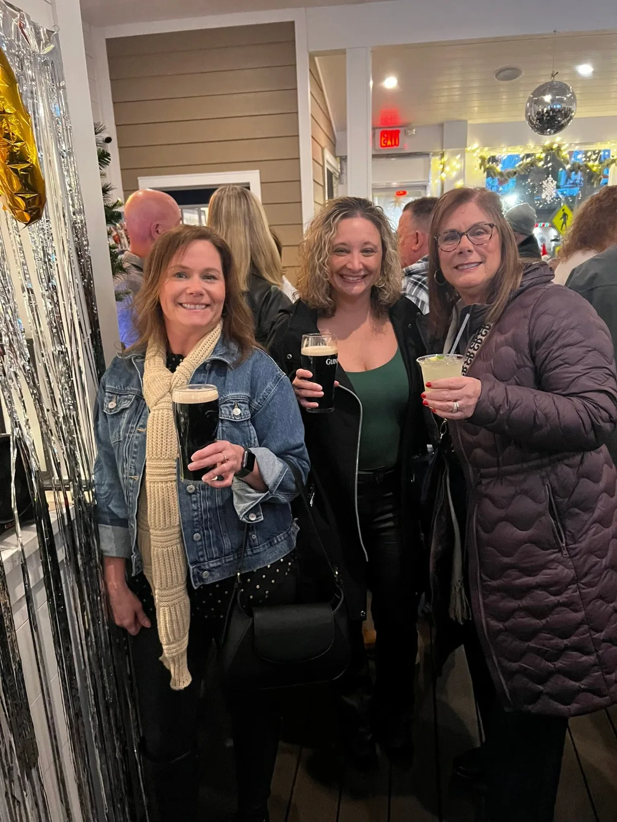 Three smiling women holding pints and cocktails stand together inside a festive, decorated bar.