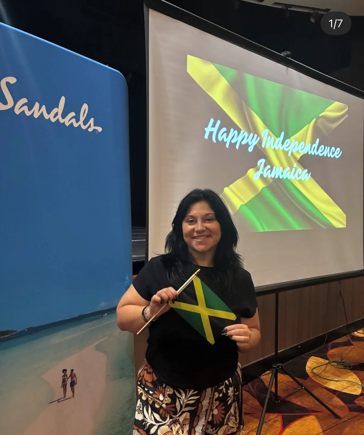 A smiling woman holds a small Jamaican flag in front of an event presentation screen and a resort banner.