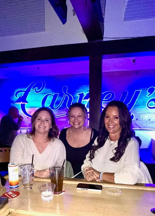 Three women smile together at a wooden bar counter with drinks, seated under vibrant blue ambient lighting.