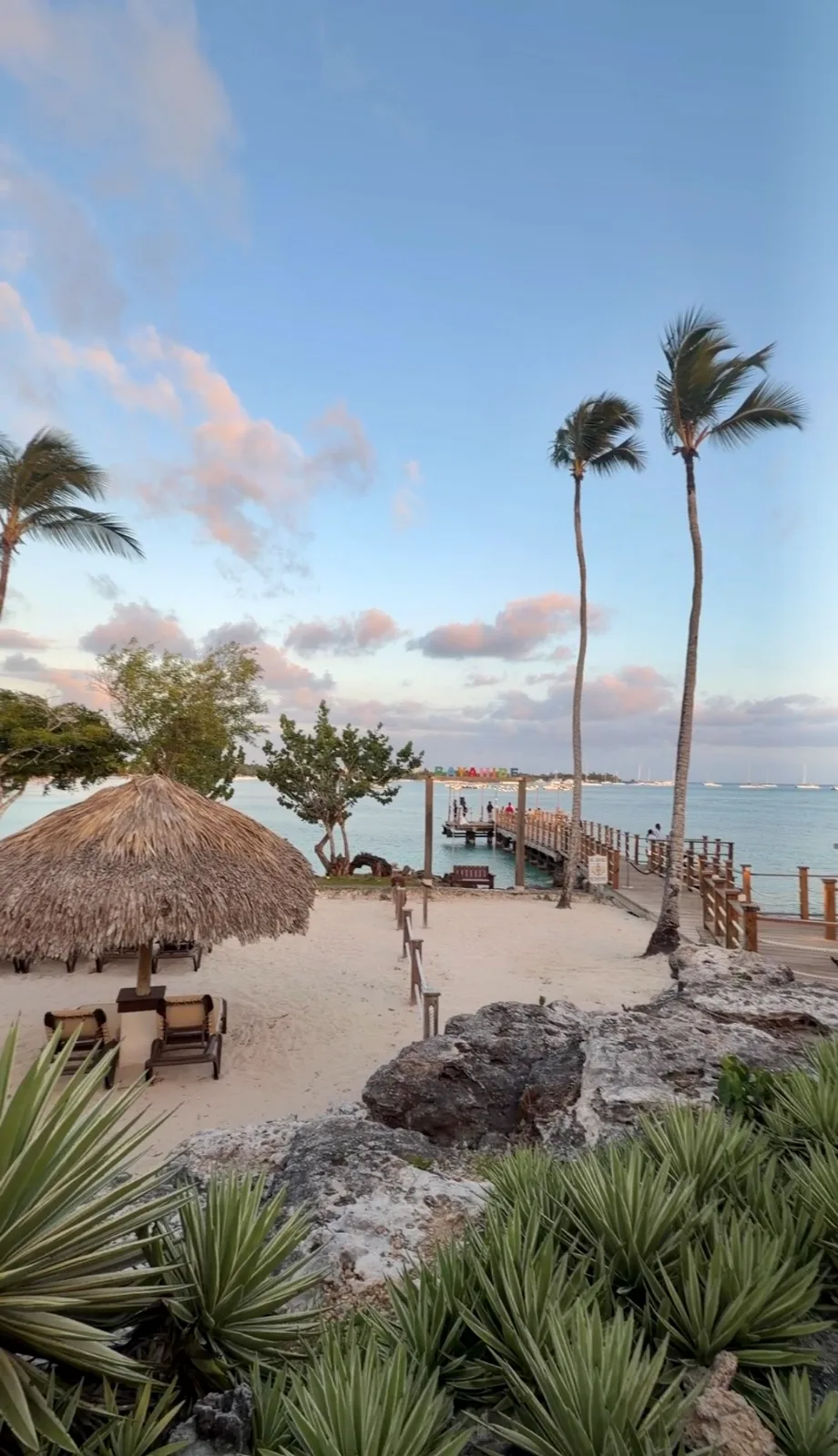 A thatched palapa and lounge chairs rest on a white sand beach beside tall palm trees and a wooden pier at dusk.