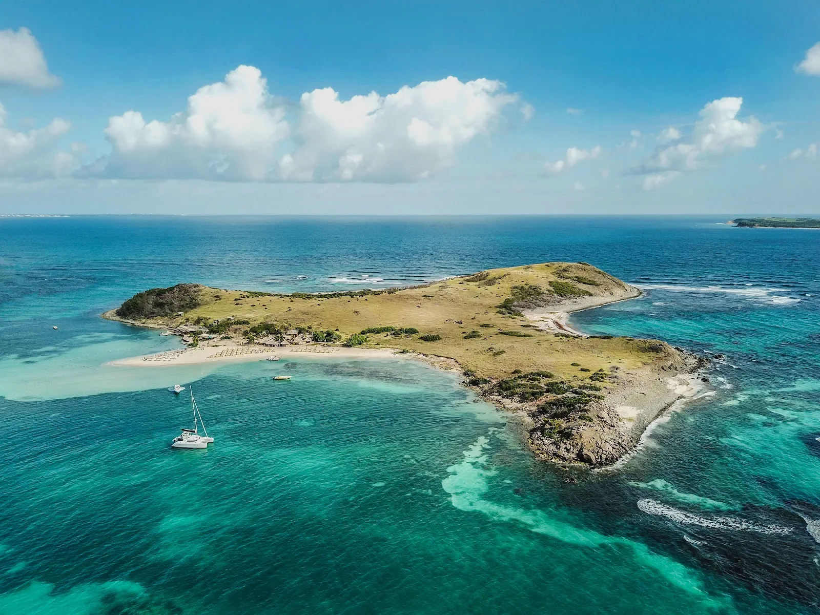 A white catamaran anchors off a sandy beach on a grassy island surrounded by vibrant turquoise Caribbean waters.