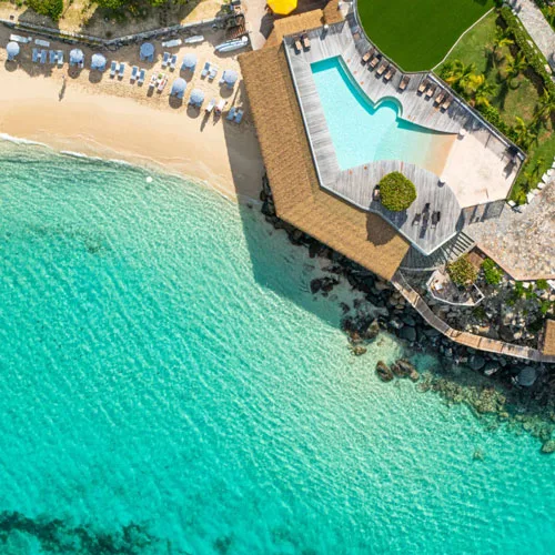 A beachfront resort pool and sun deck border a sandy bay and clear turquoise waters in St. Martin.