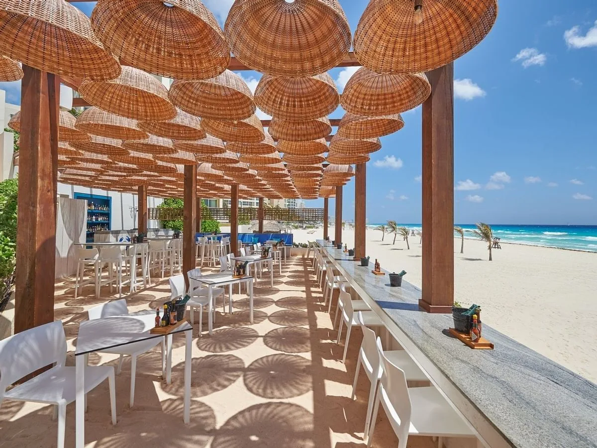 Woven wicker lanterns shade an open-air beachfront restaurant patio overlooking white sand and turquoise ocean waters in Cancún, Mexico.