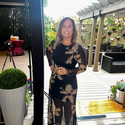 A smiling woman in a black floral dress holds a drink on a wooden outdoor terrace with pergola and lounge seating.