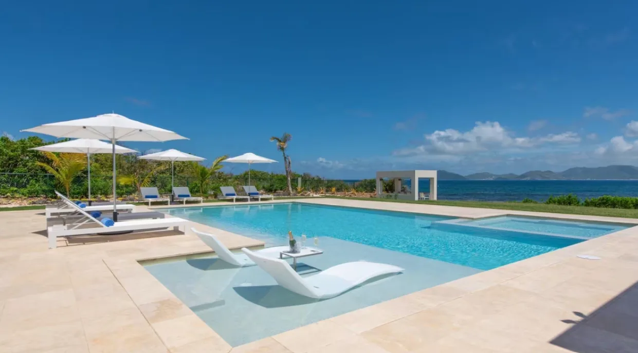 Submerged sun loungers and umbrella-shaded deck chairs line a villa swimming pool overlooking the ocean under a clear blue sky.