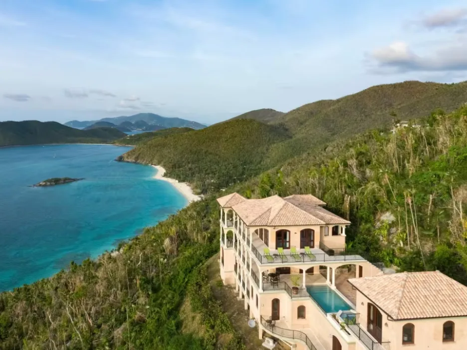 A multi-story villa with a swimming pool stands on a lush hillside overlooking a tropical turquoise bay and sandy beach.