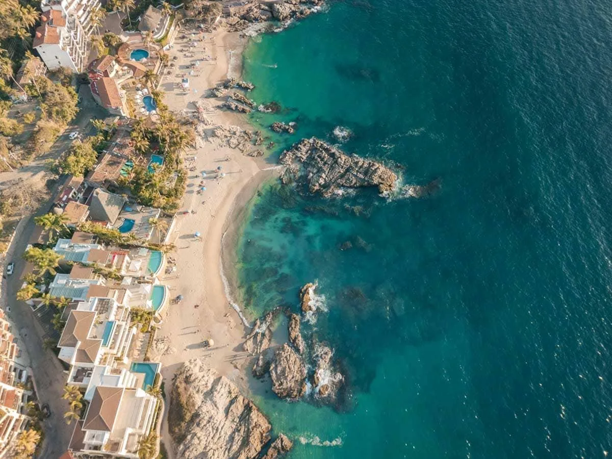 Coastal villas line a sandy beach cove surrounded by rocky outcrops and turquoise ocean water in Puerto Vallarta, Mexico.
