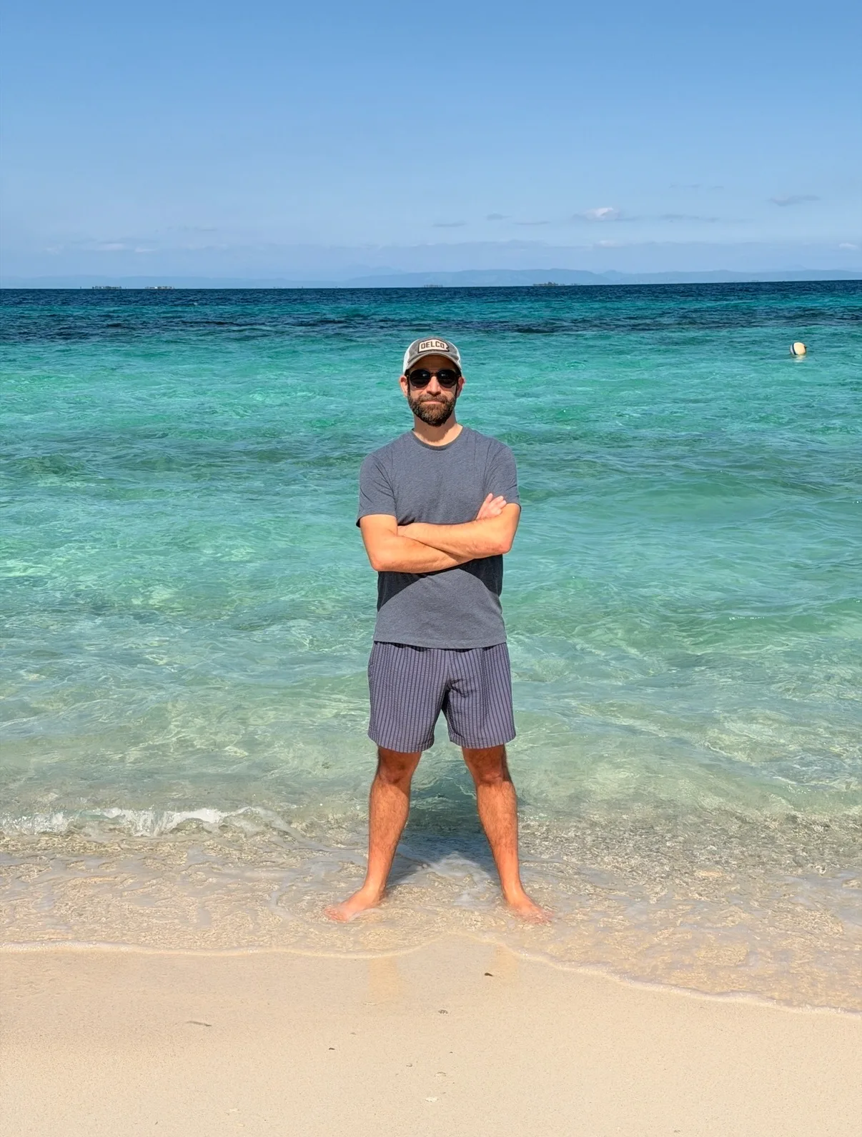 A man stands with arms crossed at the shore of a sunny beach with clear turquoise ocean water.