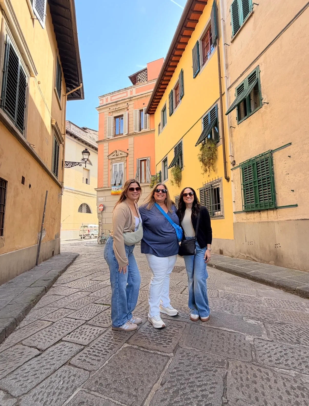 Three women smile together on a paved stone street framed by classic yellow buildings with green shutters.