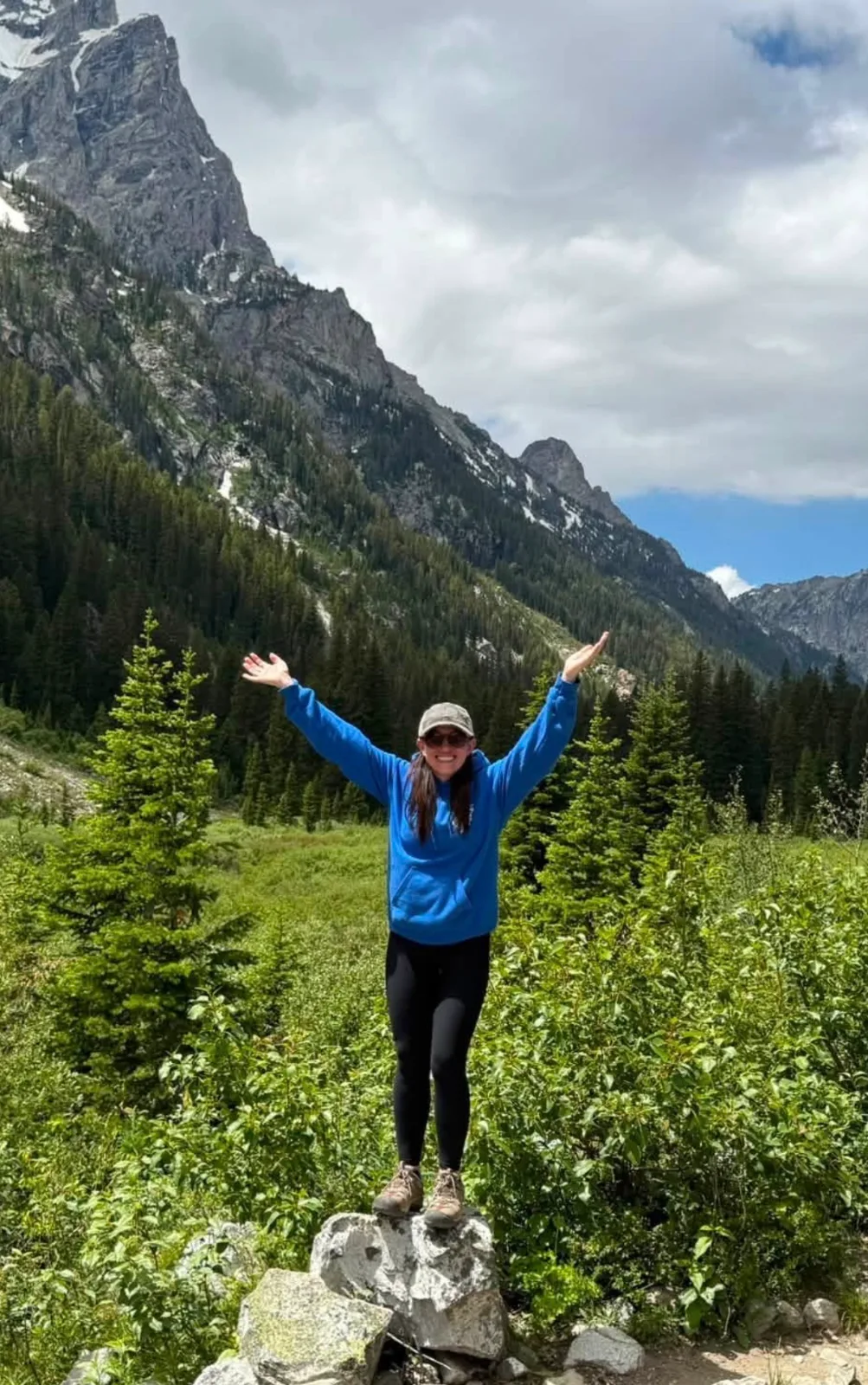 A hiker stands on a rock with open arms against a scenic backdrop of pine trees and towering mountain peaks.