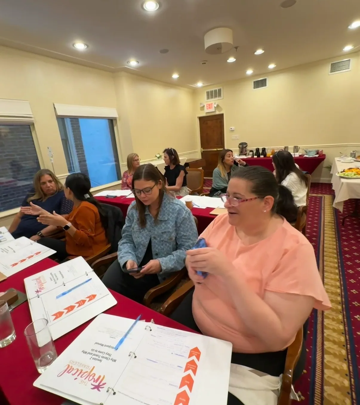 Women participate in a training session, seated at tables with open participant binders inside a conference room.
