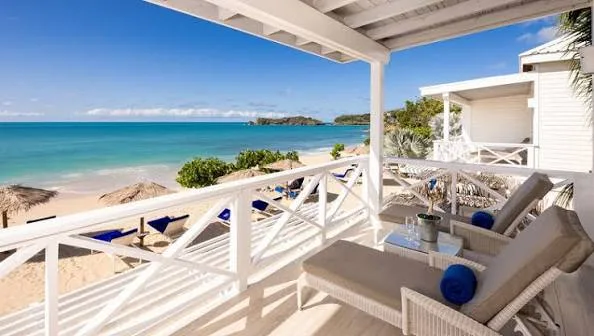 Cushioned wicker loungers rest on a white wooden balcony overlooking a sandy tropical beach and turquoise ocean in Antigua.