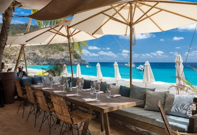 A set dining table under umbrellas at a beachfront restaurant overlooking turquoise ocean waters in St. Barthelemy.