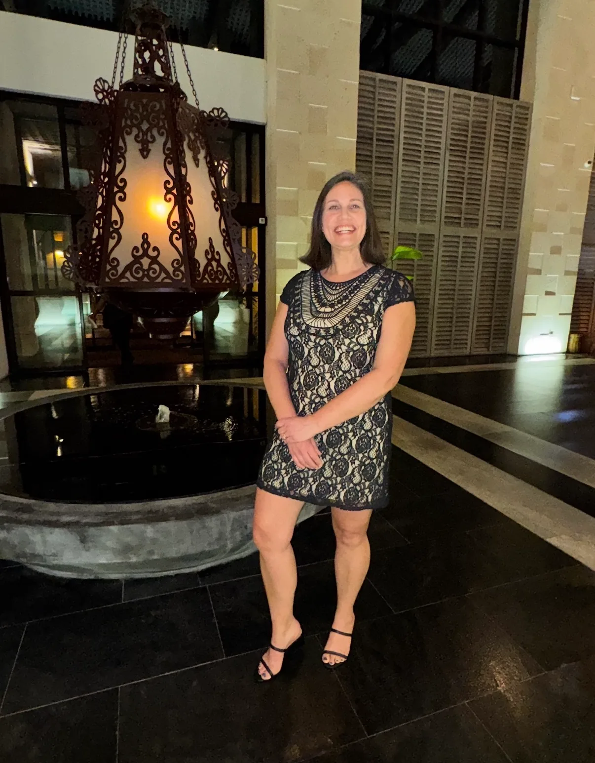 A smiling woman in a lace dress stands beside a stone fountain and an ornate hanging lantern in a hotel courtyard.