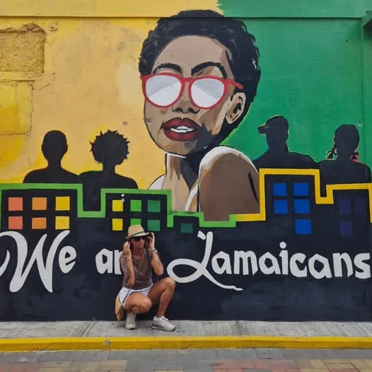 A woman in a sun hat crouches in front of a vibrant Jamaican street mural painted on an urban wall.