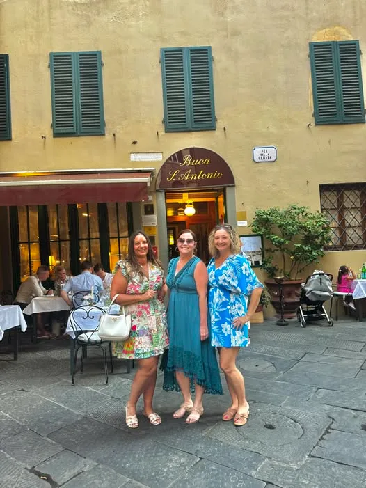 Three women in colorful summer dresses stand outside a traditional restaurant with outdoor dining tables on a paved street.