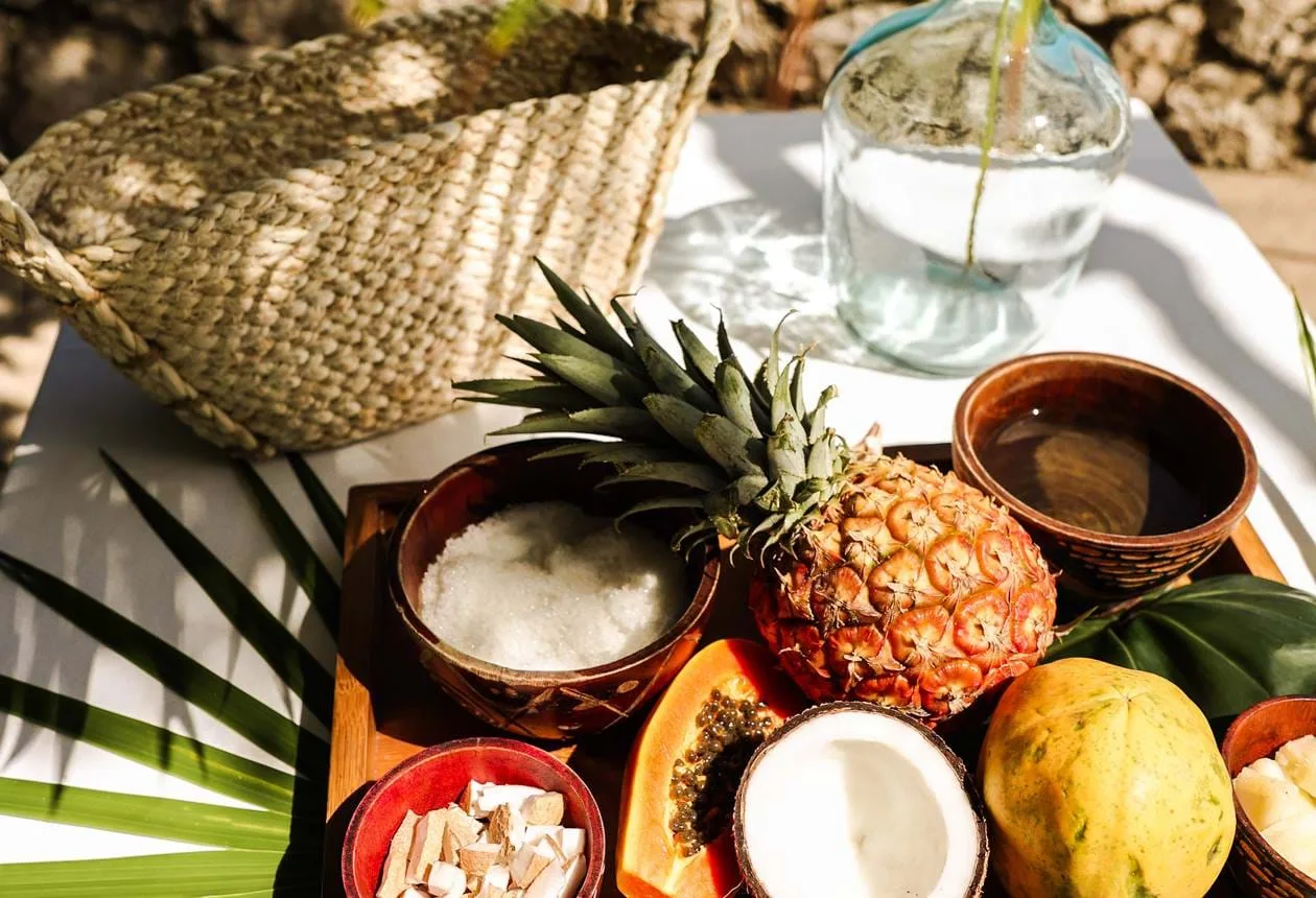 Fresh tropical fruits, coconut, and bowls of natural spa ingredients arranged on a sunlit table beside a woven basket.