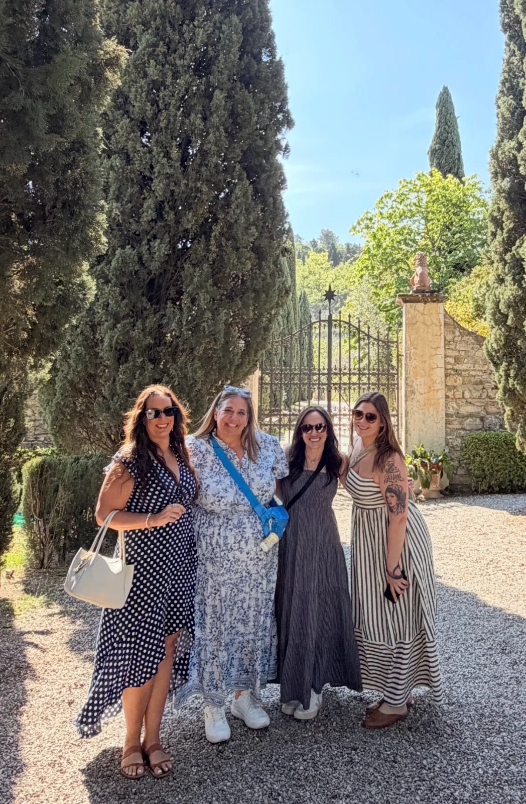 Four women in maxi dresses smile together on a gravel driveway before tall cypress trees and an ornate estate gate.