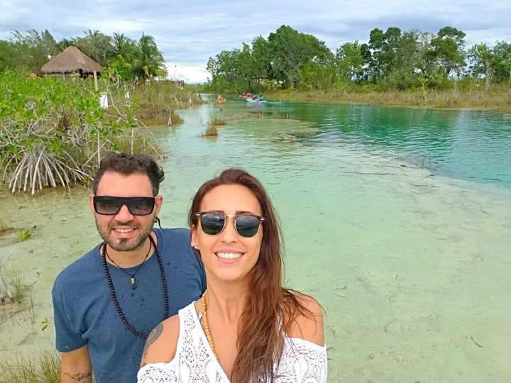 A smiling couple in sunglasses poses before a clear turquoise waterway lined with lush mangroves and tropical greenery.