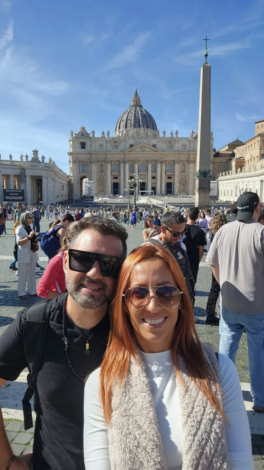 A smiling couple wearing sunglasses takes a selfie in front of St. Peter's Basilica and the obelisk in Vatican City.