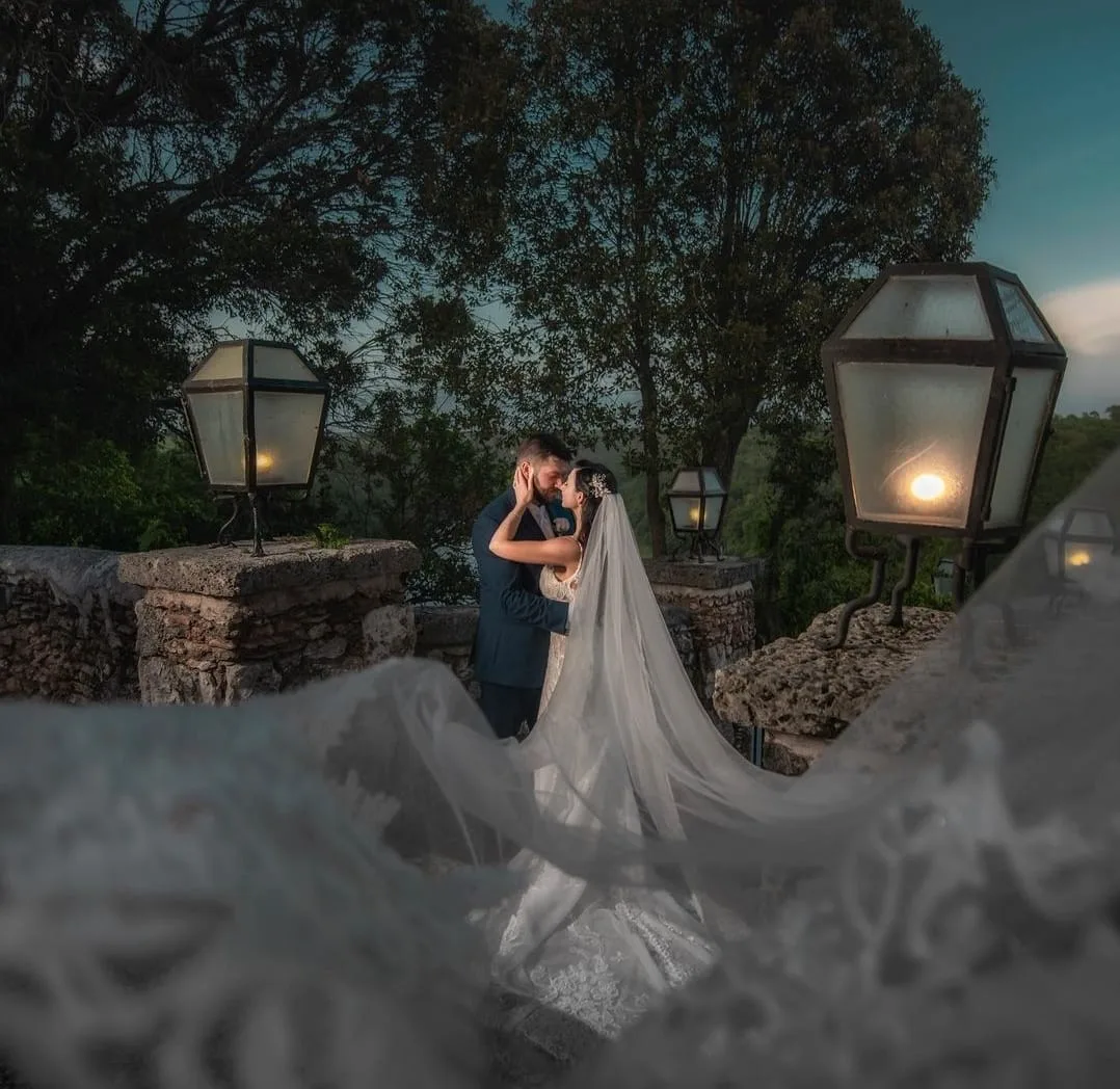 A bride and groom embrace on a stone balcony framed by illuminated vintage lanterns and a flowing veil at dusk.