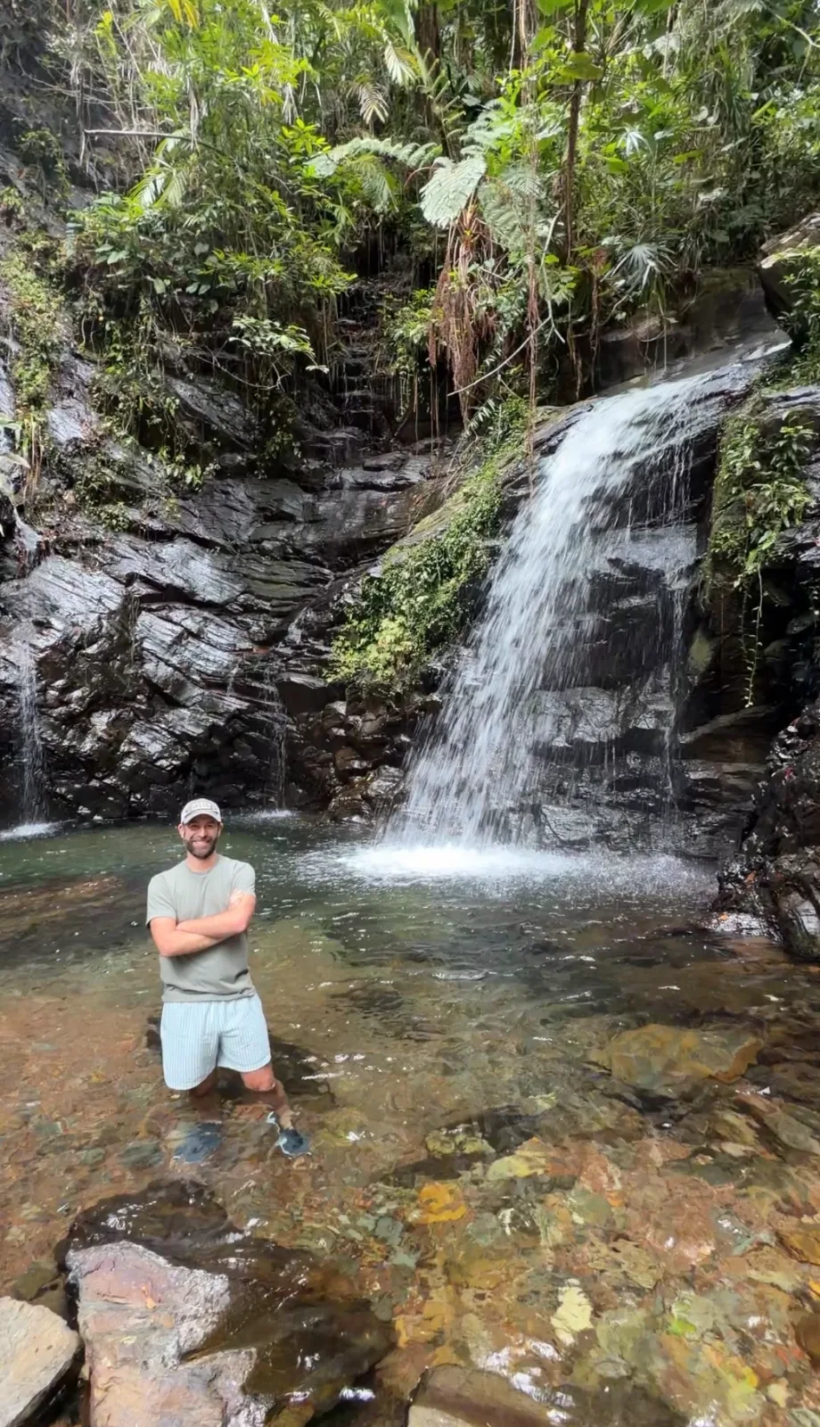 A smiling man stands in a clear natural pool below a cascading waterfall surrounded by dense tropical foliage.