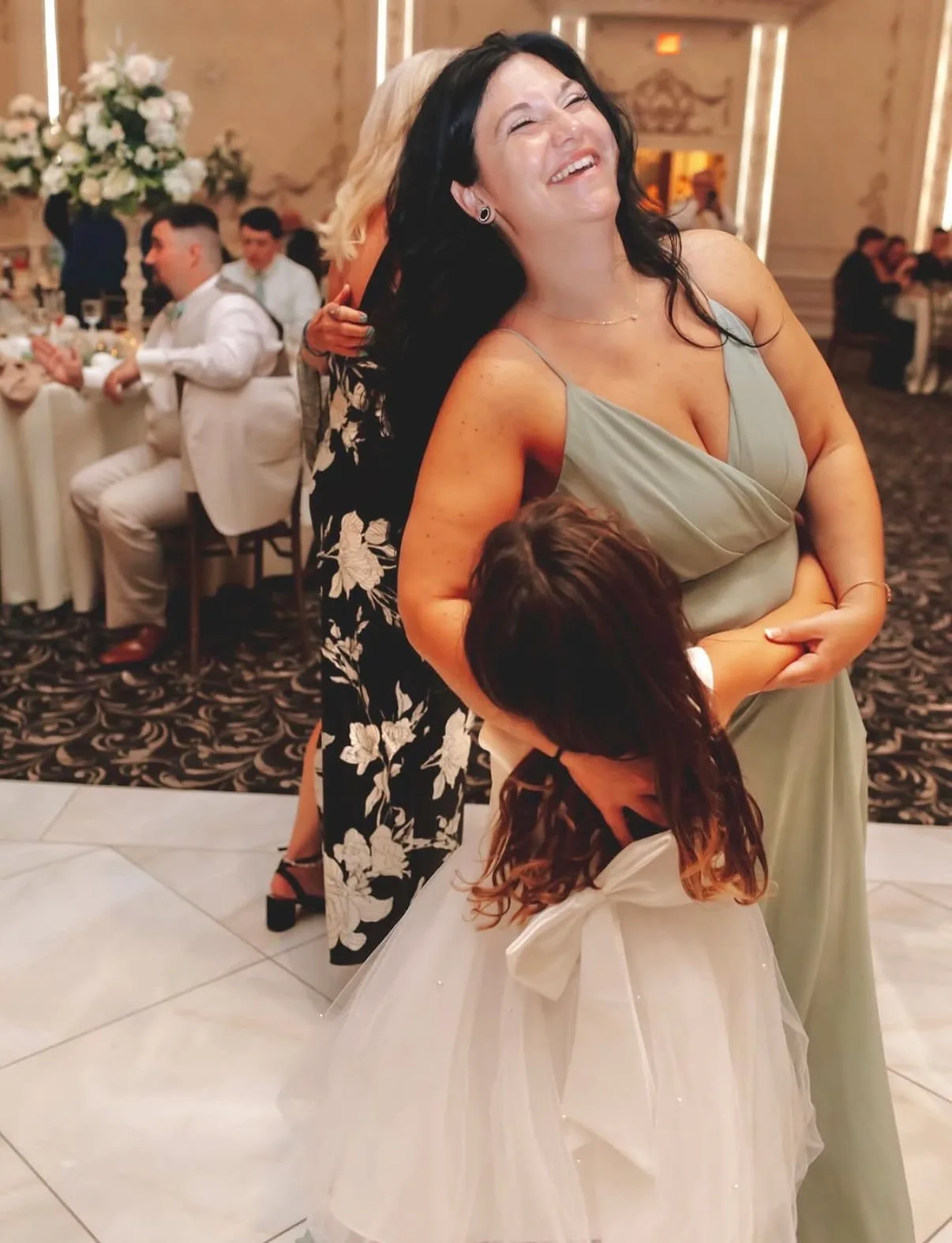 A laughing woman in a sage green dress dances with a young flower girl inside an elegant banquet ballroom.
