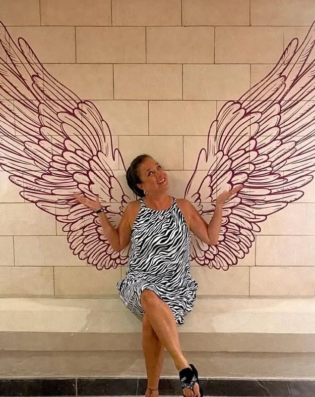 A smiling woman in a zebra-print dress poses with open arms against a wall painted with large angel wings.