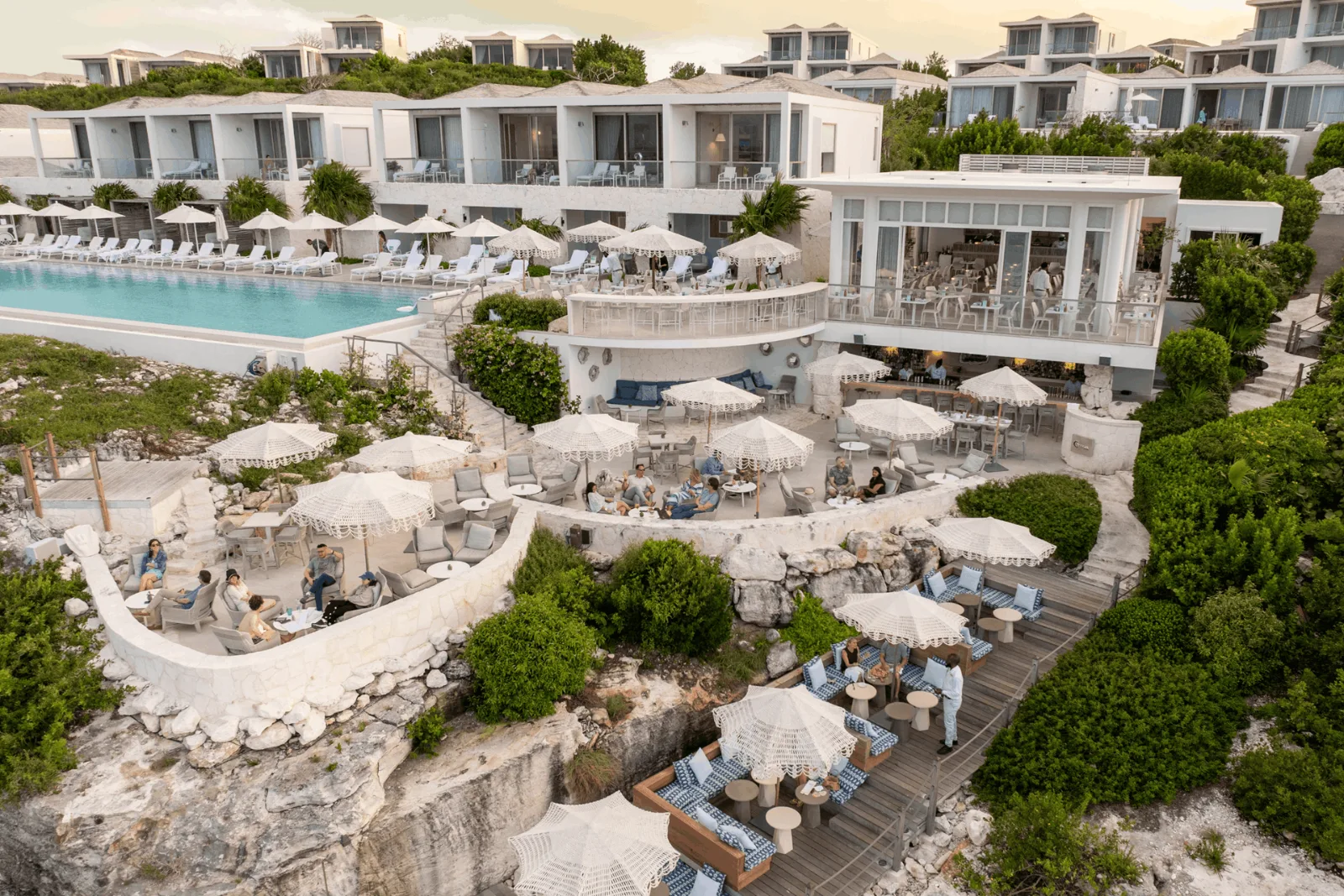 Tiered cliffside lounge terraces with fringed umbrellas descend below a modern white resort and swimming pool.