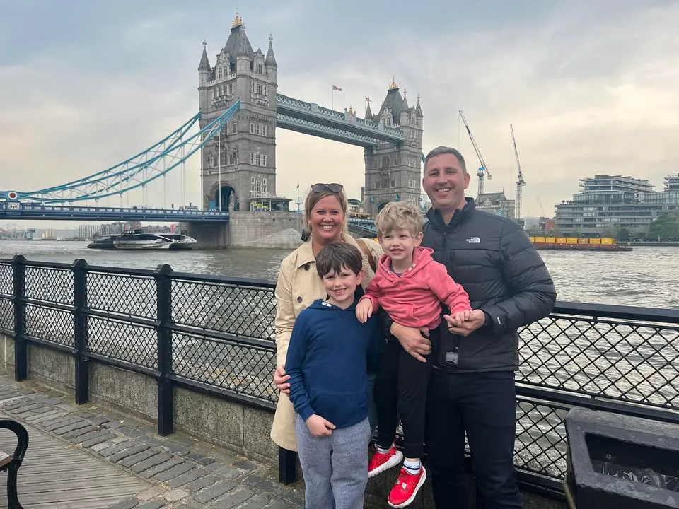 A smiling family of four stands along the River Thames promenade with London's Tower Bridge in the background.