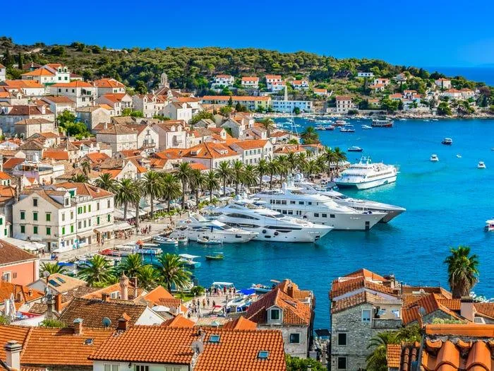 Luxury yachts dock along a palm-lined harbor surrounded by terracotta-roofed stone buildings in Hvar, Croatia.