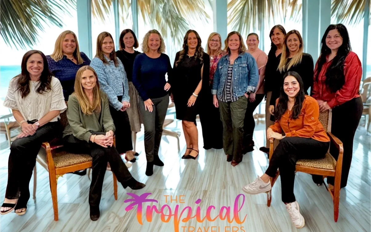 A group of smiling women poses together in a bright room lined with large windows overlooking palm trees.