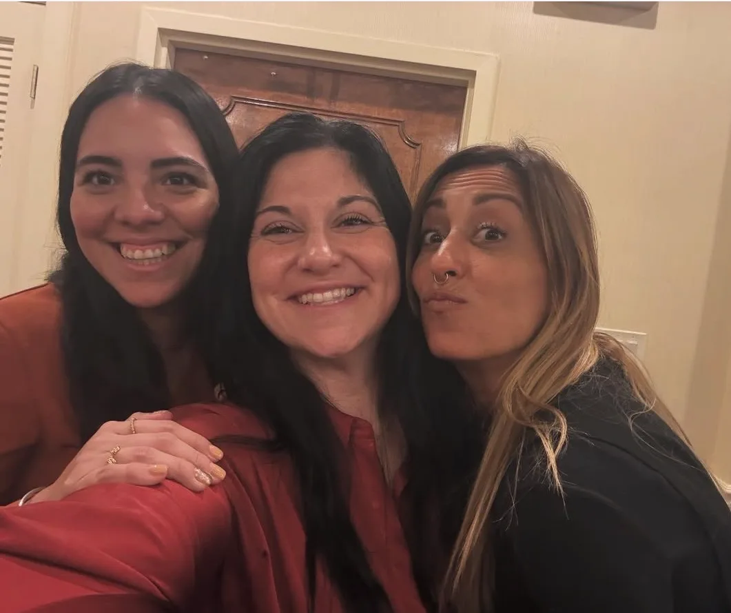 Three smiling women pose closely together for a selfie indoors in front of a wooden door.