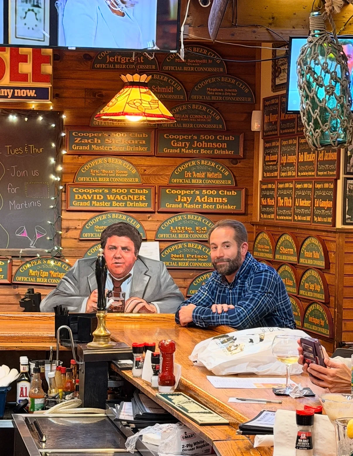 A man in a plaid shirt sits beside a cardboard cutout at a rustic wooden bar decorated with beer club plaques.