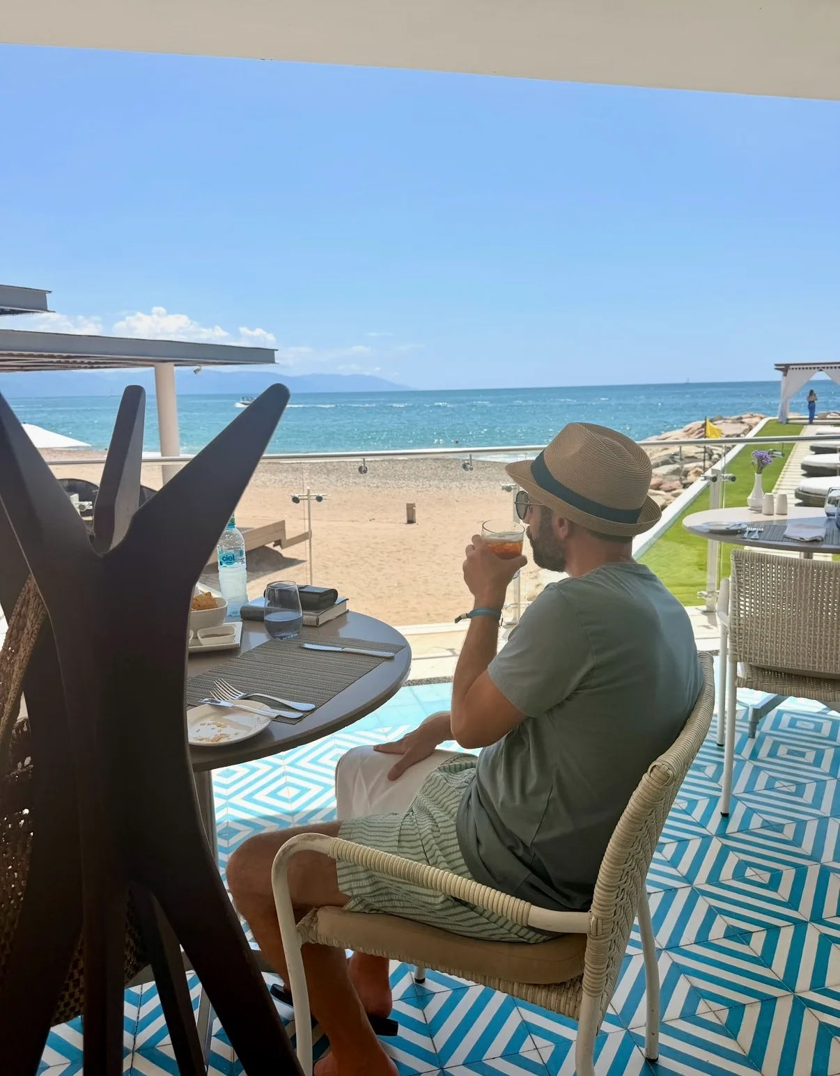 A man in a straw hat drinks at an outdoor restaurant table overlooking a sandy beach and ocean.