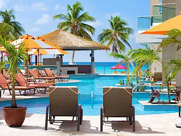 Lounge chairs line a resort swimming pool overlooking a thatched pavilion, palm trees, and the ocean in Barbados.