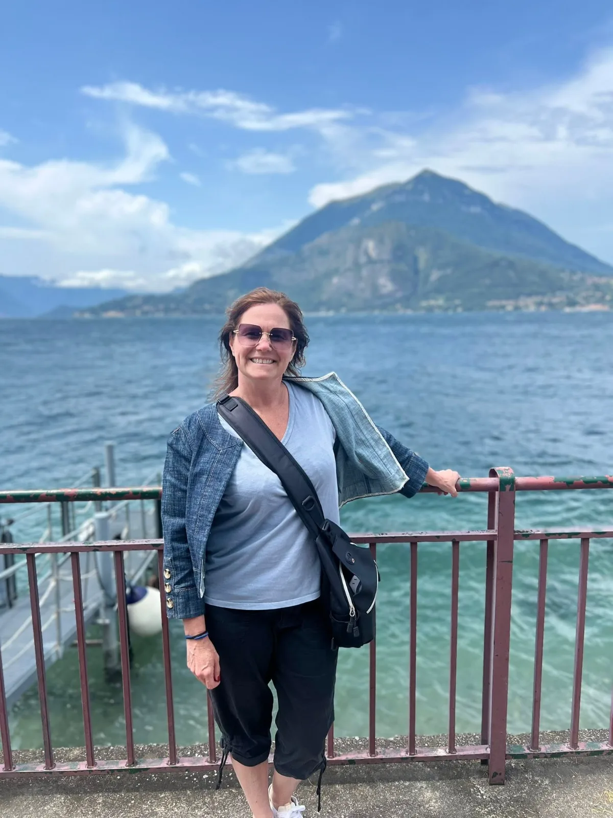 A smiling woman wearing sunglasses stands beside a waterfront railing with a vast lake and distant mountains behind her.