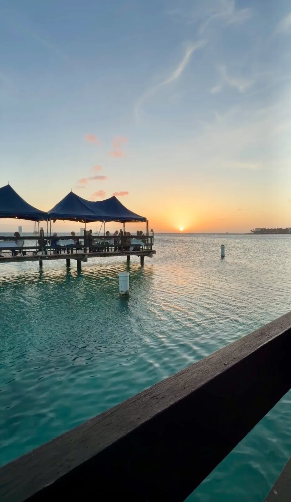 Diners sit beneath shaded canopies on a wooden pier over calm turquoise ocean waters at sunset in Aruba.