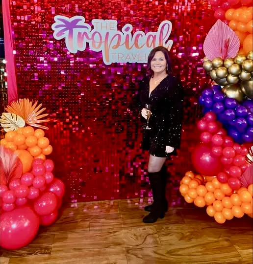 A smiling woman in a black sequin dress poses before a shimmering red backdrop flanked by colorful balloon arrangements.