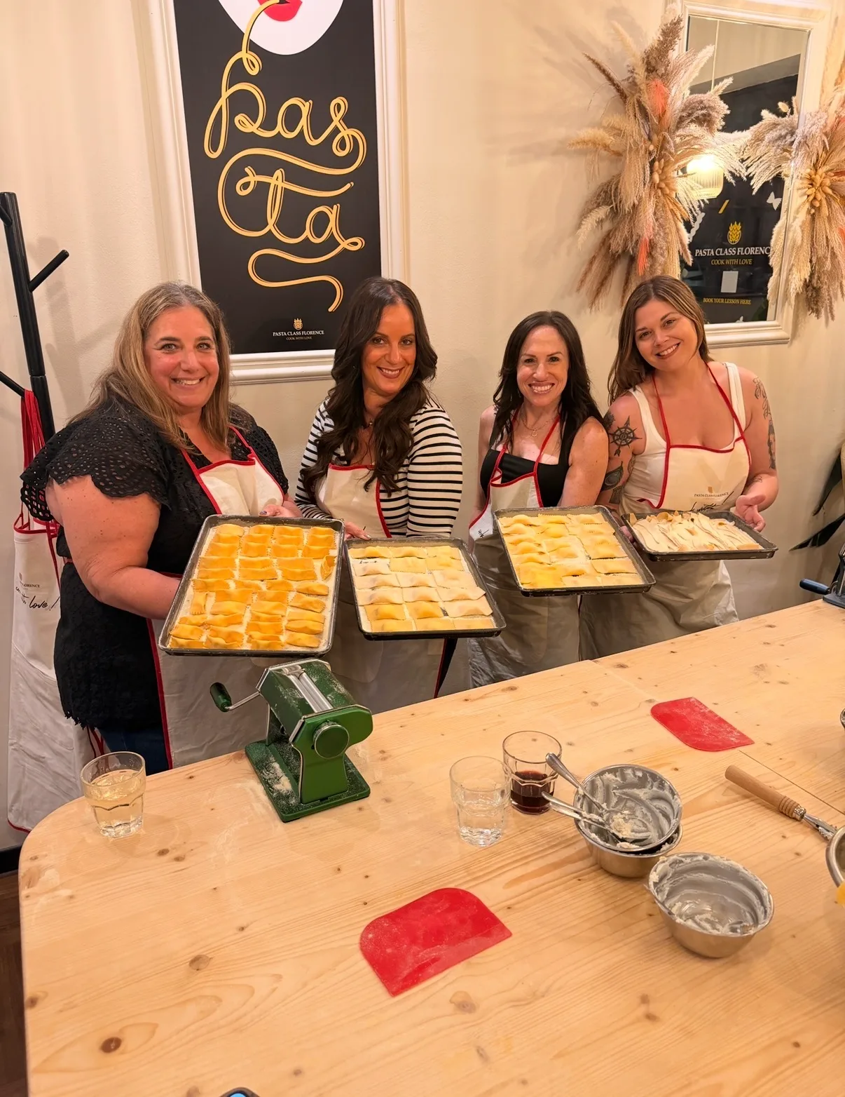 Four smiling women in aprons hold trays of handmade ravioli behind a wooden prep table during a cooking class in Florence.