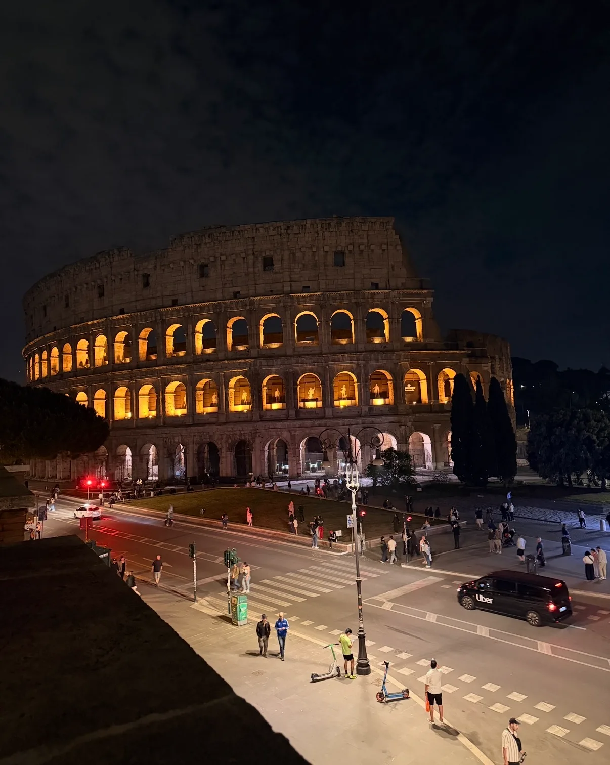 The illuminated Roman Colosseum glows at night above a city street with pedestrians and passing vehicles in Rome, Italy.