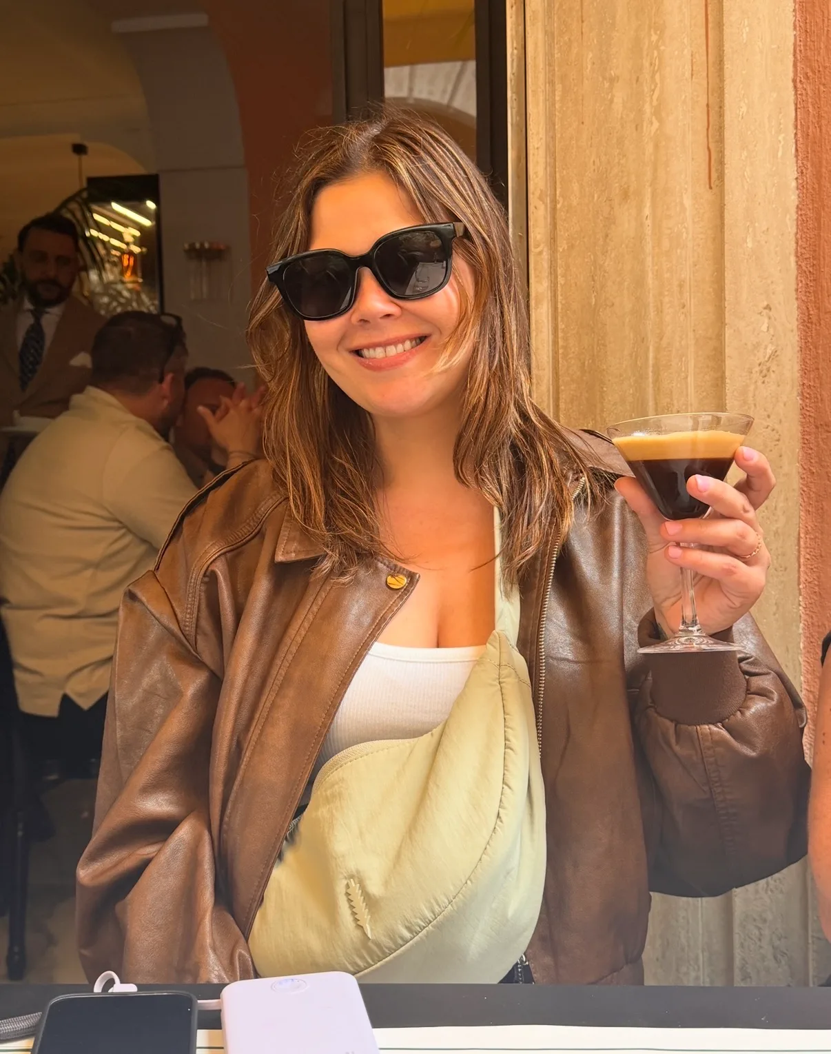 A smiling woman wearing sunglasses and a brown leather jacket holds an espresso martini at an outdoor cafe table.