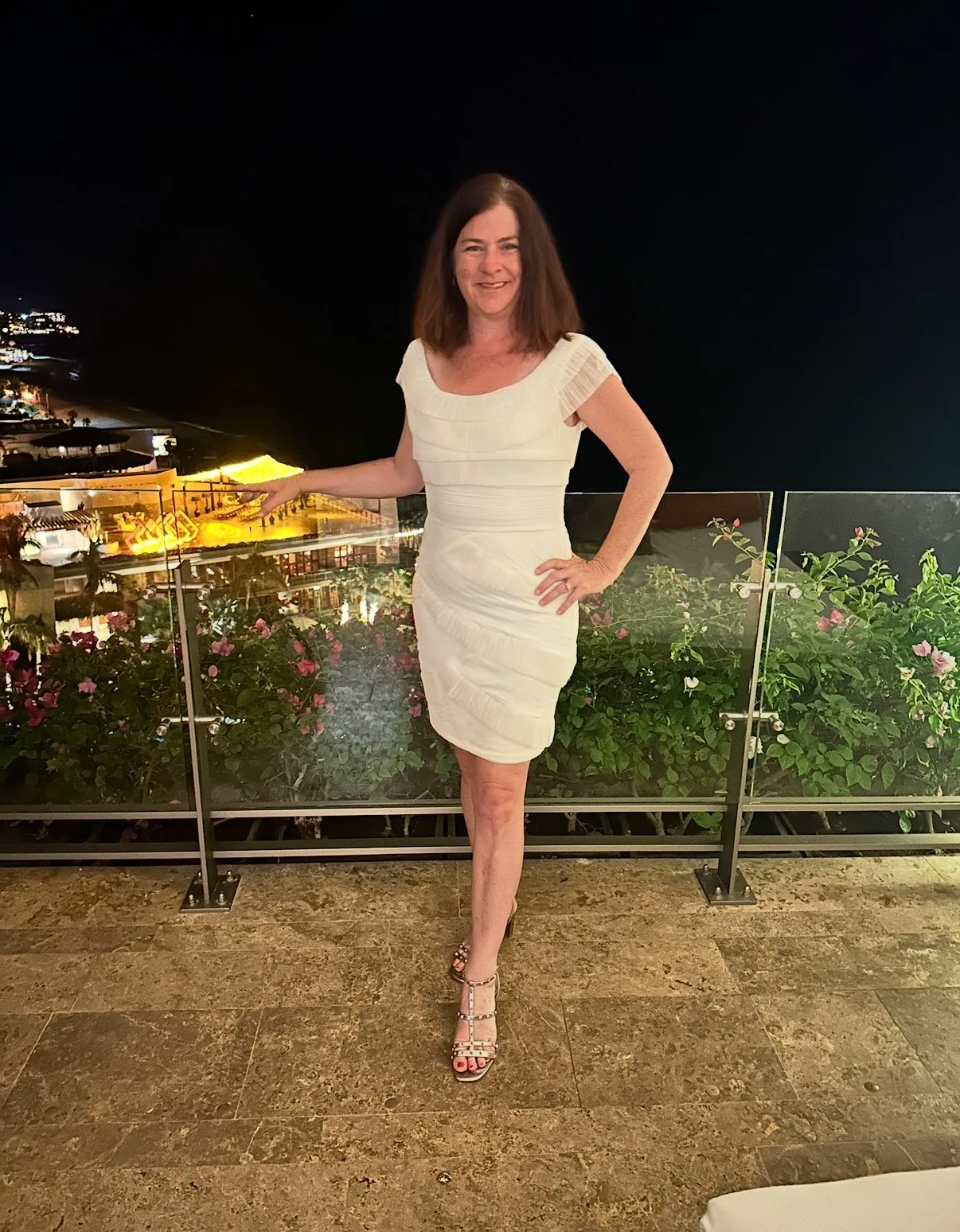 A smiling woman in a white dress poses on a terrace balcony overlooking an illuminated resort at night.