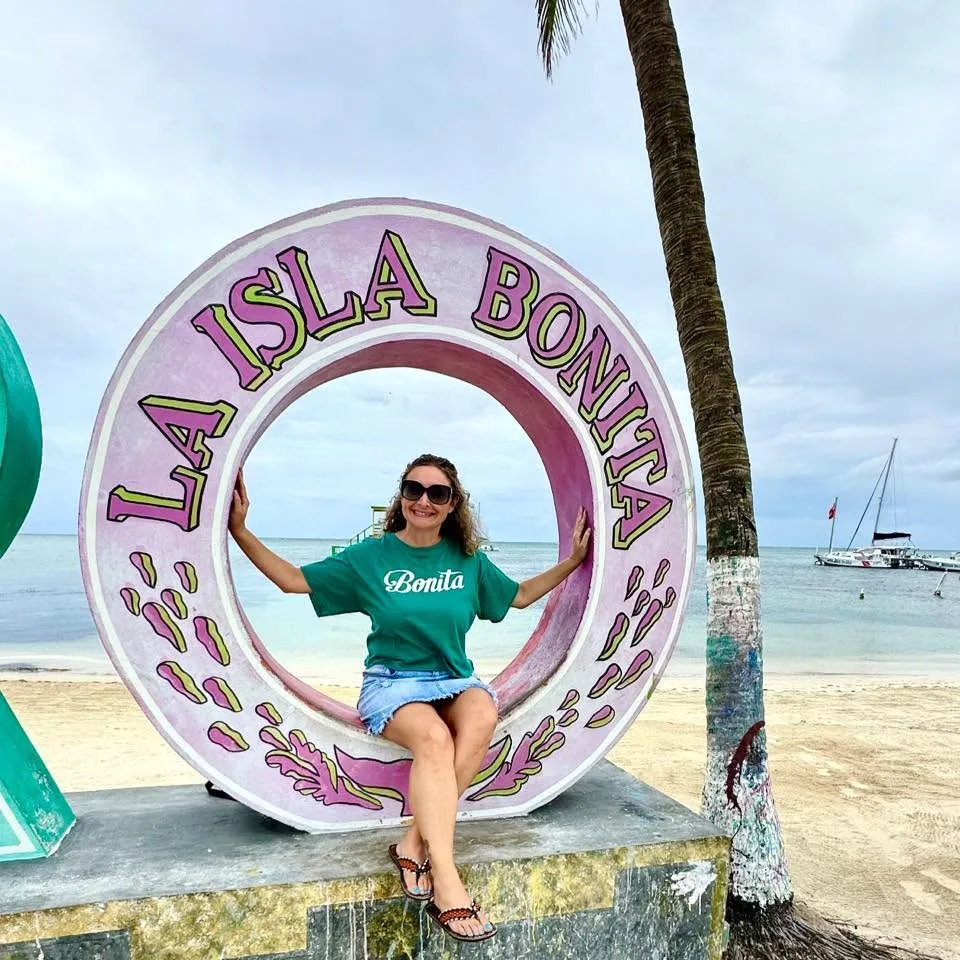 A smiling woman sits inside a circular La Isla Bonita sign on a sandy beach beside a palm tree and calm sea.